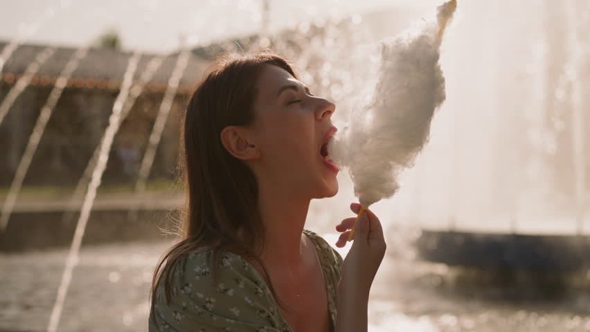Pretty lady eats sugar cotton wool against blurry fountain at sunset time closeup slow motion. Street dessert in urban park. Woman remembers carefree childhood Royalty-Free Stock Footage #1103690923