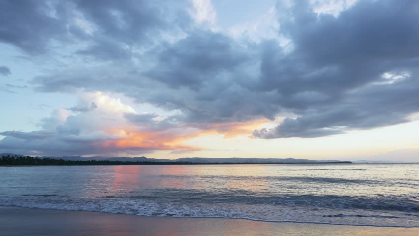 Landscape of the morning sky with a sun glow in thick clouds over the ocean. Calm sea waves on a sandy beach at dawn. Exotic nature view. Inspirational seascape. Twilight on the beach.