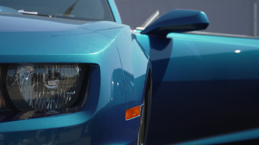 Blue American muscle car is parked on sun with door open and parking fines under windshield wiper blade. Headlight and orange marker on clean polished metallic bodywork.