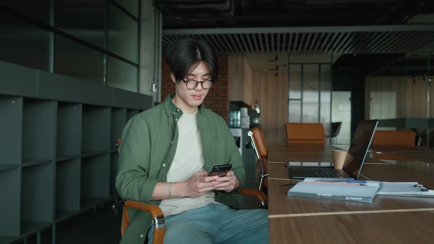 Smiling Asian man in eyeglasses typing on mobile in a co-working room Royalty-Free Stock Footage #1103707017