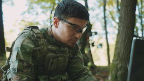 Close-up portrait of soldier in camouflage clothes using laptop computer and looking focused on screen in forest. Blurred view of two soldiers holding drone on background. Surveillance and defense - Powered by Shutterstock - Get 15% off with code: PIKWIZARD15