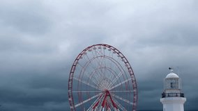 A dramatic and moody video of a spinning ferris wheel against a dark stormy sky, with the top of a lighthouse building and a seagull flying around