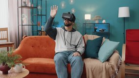Excited man using virtual reality futuristic technology VR app headset helmet to play simulation 3D 360 video game watching film movie. Portrait of guy in goggles at modern home apartment room on sofa