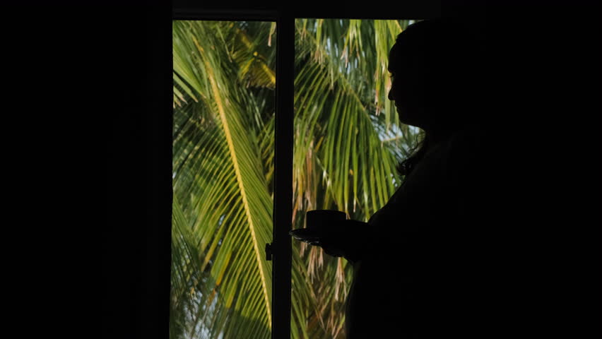 Young woman drinking coffee by the window with tropical view, slow motion. Palm leaves swaying outside Royalty-Free Stock Footage #1103714345