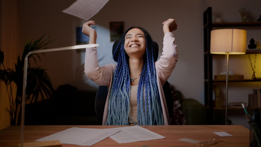 Delightful happy African American young woman journalist, reading document on paper sheet, throwing articles and feeling excited and happy by successful work, sitting at wooden desk at home in night