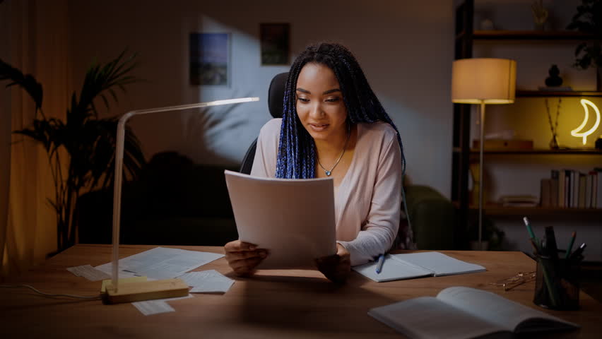 Happy young African American stylish student, expressing happy positive emotions while reading article, sitting at wooden desk in the cozy home interior. People. Education. Career. Lifestyle concept