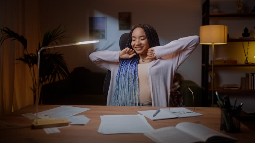 Authentic portrait of happy female student journalist, experiencing satisfaction by her article, sitting at wooden desk in the cozy home interior and reading her essay in the night time. People Career