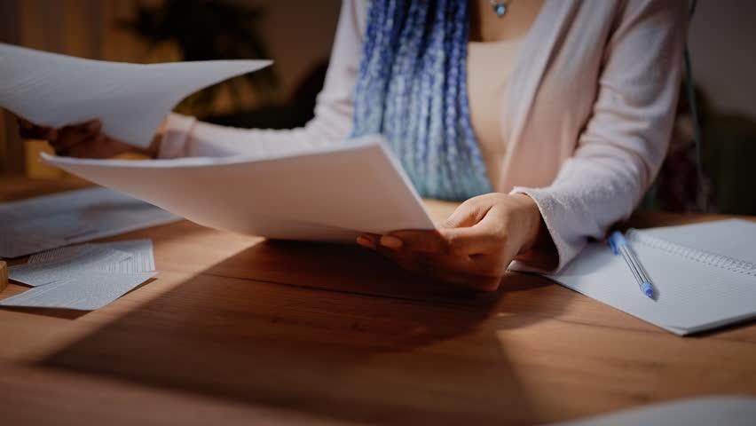 Close-up woman's hands flipping through sheets of paper while sitting at a desk. Cropped view of journalist reviewing her article for magazine or young student analyzing her essay or graduation work