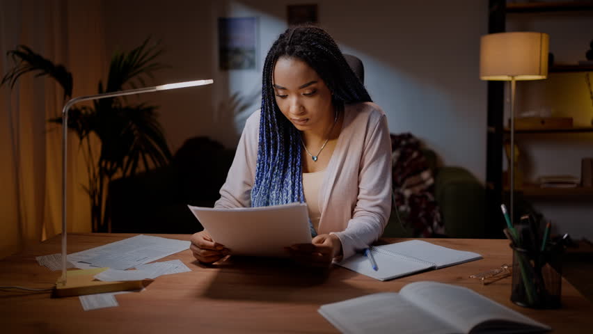 Portrait of confident focused African American young woman journalist, flipping through sheets of paper while reading and reviewing her article or document, sitting at a desk in the night time.