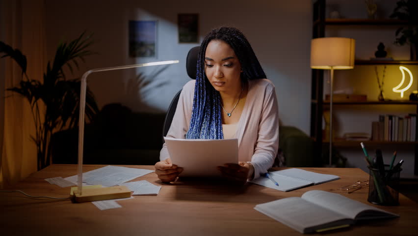 Confident African American young woman journalist sitting at desk in the night time and reading her new article for a journal or magazine. Determined university graduated student reviewing her essay