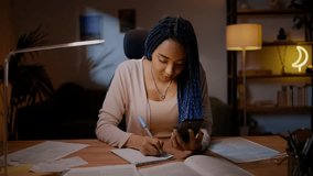 African American young confident student writing essay, holding mobile phone in her hands, working on graduation project in the night time, sitting at desk in the living room. People Education Career - Powered by Shutterstock - Get 15% off with code: PIKWIZARD15