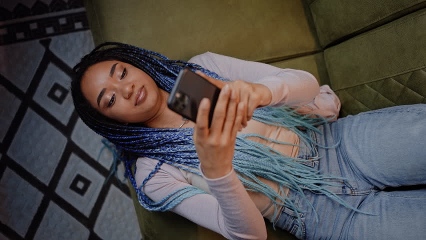 Top view African pretty woman with stylish dreadlocks, using mobile phone, lying on sofa at home, writing sms, checking social media content, communication with friends via internet chat. Slow motion Royalty-Free Stock Footage #1103716247