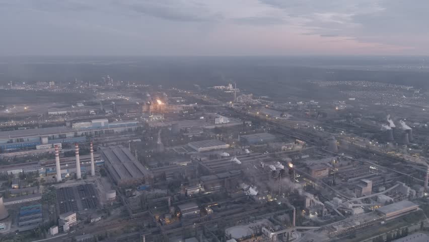 Lipetsk, Russia. An extensive iron and steel works complex sprawls across the Left Bank District in a sweeping aerial view captured after sunset. The massive industrial site is defined by multiple tall chimneys releasing plumes of smoke and steam, large factory halls with illuminated roofs, cooling towers, and an intricate network of pipelines and railway tracks that weave between the structures. The scene is set during the blue hour, with the sky retaining faint traces of twilight while artificial lights from the plant and surrounding infrastructure begin to dominate the landscape. The overall atmosphere is one of heavy industry and continuous operation, with the warm glow of furnaces and torch flames contrasting against the cool dusk tones. The camera maintains a steady, elevated perspective that emphasizes the vast scale and dense layout of the metallurgical facility. A-Log.