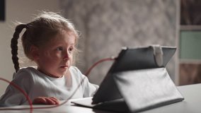 Emotional upset little girl with plait watches video lesson via tablet PC sitting at table in light children room close view slow motion