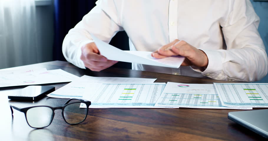 Investor inspects paper documents with data in spreadsheets. Office employee in white shirt sits at wooden table with glasses and mobile phone Royalty-Free Stock Footage #1103729855