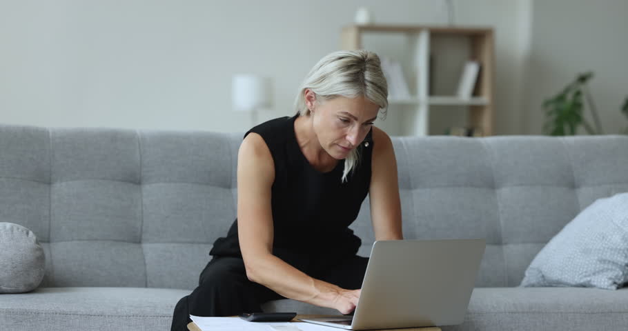 Serious mature homeowner woman using financial application on laptop, counting budget, reviewing paper bills, invoices, paying taxes, insurance fees. Freelancer accountant working at home