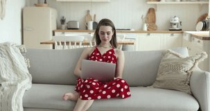 Positive young adult gen Z girl in red cloth typing on laptop at home, using Internet service, app, software, browsing website, watching online video, smiling, laughing, looking away