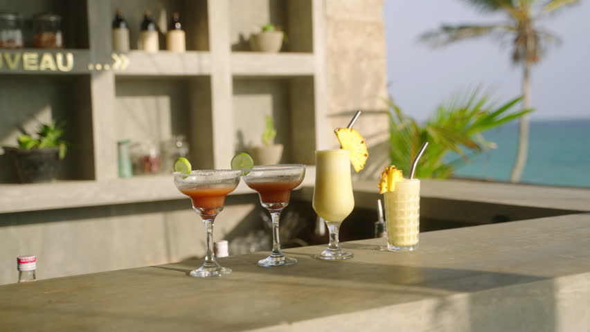 Tropical cocktails on bar at seaside restaurant. Glasses with refreshing alcoholic drinks stand on bar counter of sea view cafe. Summer open air bar. Outdoor party celebration, welcome beverage. Royalty-Free Stock Footage #1103750545