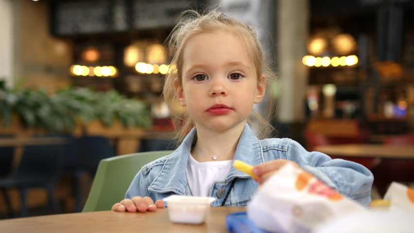 Girl eats French fries, dips potatoes in red ketchup sauce, puts food in his mouth and chews. Portrait blond Caucasian girl in denim jacket sitting at table in fast food restaurant and eating food. Royalty-Free Stock Footage #1103753981