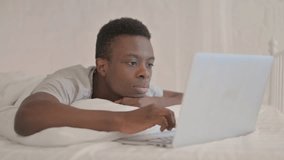 Young African Man Doing Video Chat on Laptop while Lying on Stomach in Bed