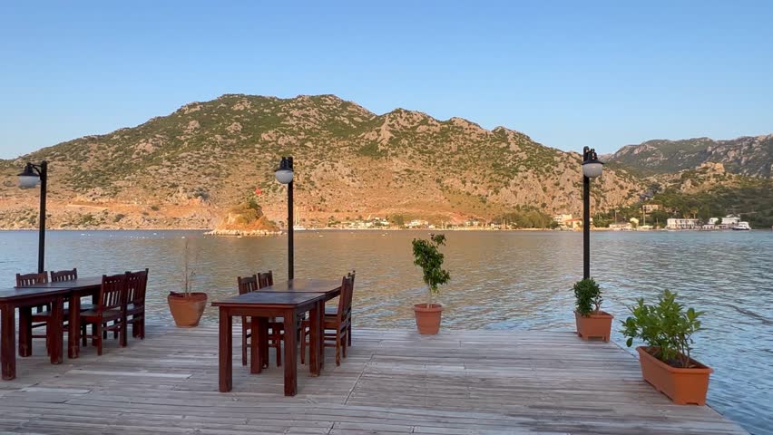 A wooden deck restaurant over sea, surrounded by tables and chairs. Calm water reflects mountains in distance. Peaceful serene scene with only sound being gentle lapping of water against shore
