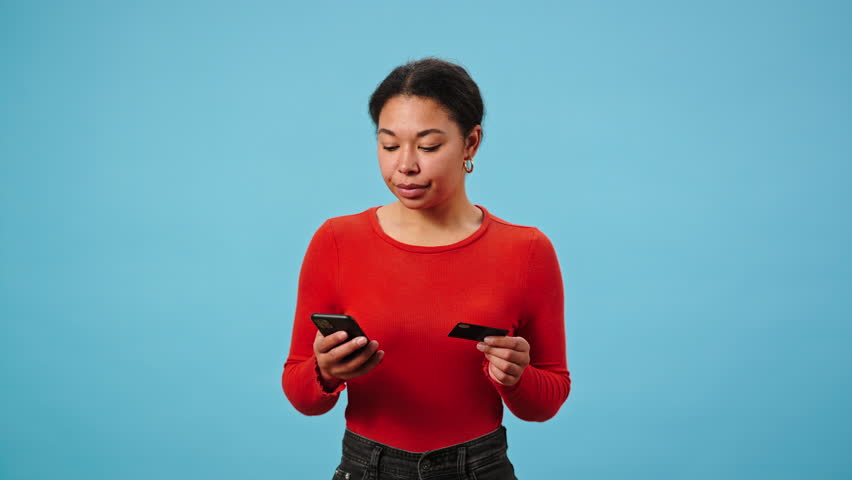 African American woman makes online purchase. Female enters credit card details and presses hands to chest looking into distance with dreamy expression in blue studio