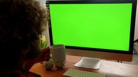 A man with curly hair sits in front of a monitor and drinks coffee from a white cup. A man works remotely at a computer while sitting in his home office.