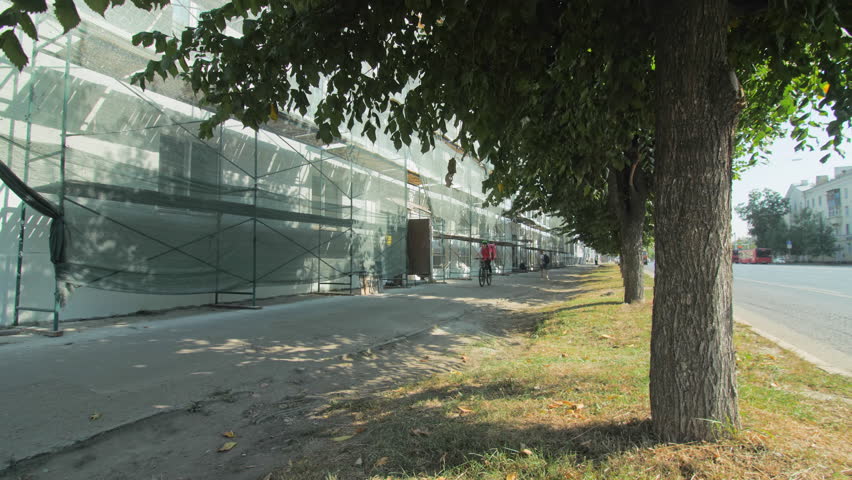 Food delivery courier rides bike past reconstructed building with scaffolds and mesh in summer city. Urban construction site and services