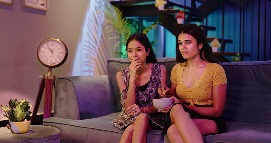 Indian two female sitting on sofa watching sad movie on TV together eat popcorn snacks late night at home. Beautiful upset women looking sentimental dramatic reality show spend weekend time  | Shutterstock HD Video #1103768463