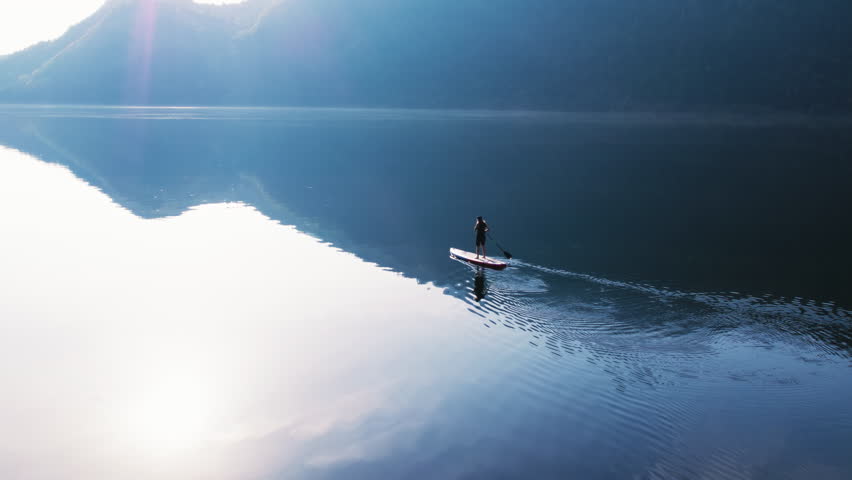 Epic and cinematic serene aerial shot of single paddle board move through still fjord water in Norway. Stand up paddle board in amazing scenery. Drone shot of paddle board in fjord Royalty-Free Stock Footage #1103774975