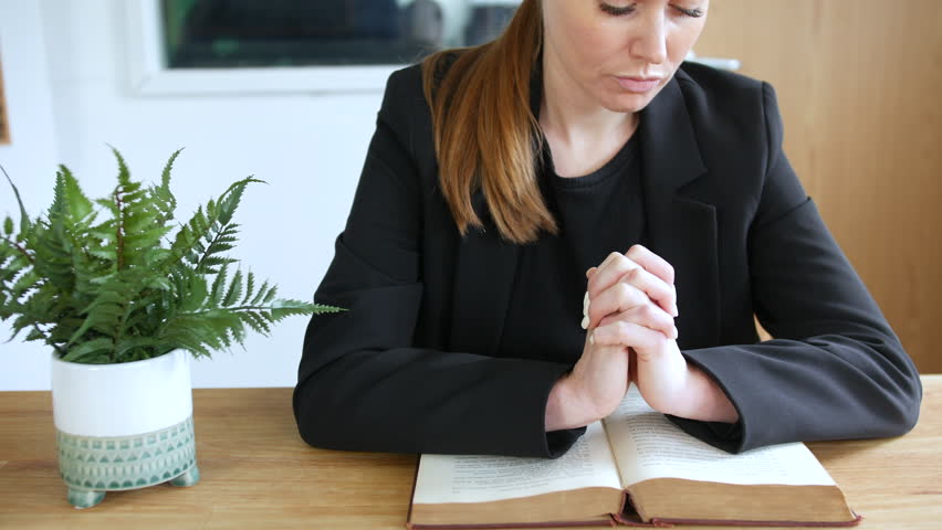 A woman prating with eyes closes and hands together in prayer over an open bible on a table