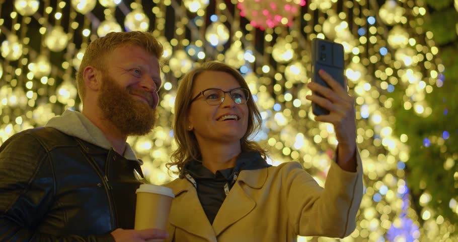 Close-up video of a loving middle aged couple during online video call with relatives or friends. They embracing on the street on the Christmas eve. Slow motion 4k footage.