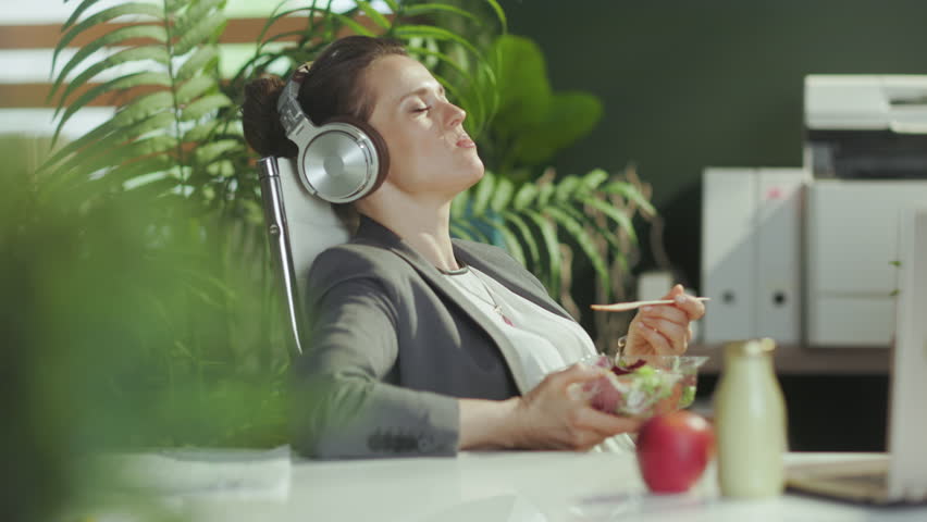 Sustainable workplace. smiling modern small business owner woman in a grey business suit in modern green office with headphones eating salad. Royalty-Free Stock Footage #1103795679