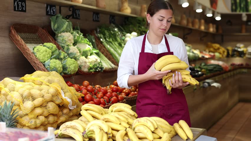 Attentive young saleswoman placing ripe bananas on food stall in grocery store . High quality 4k footage Royalty-Free Stock Footage #1103796115