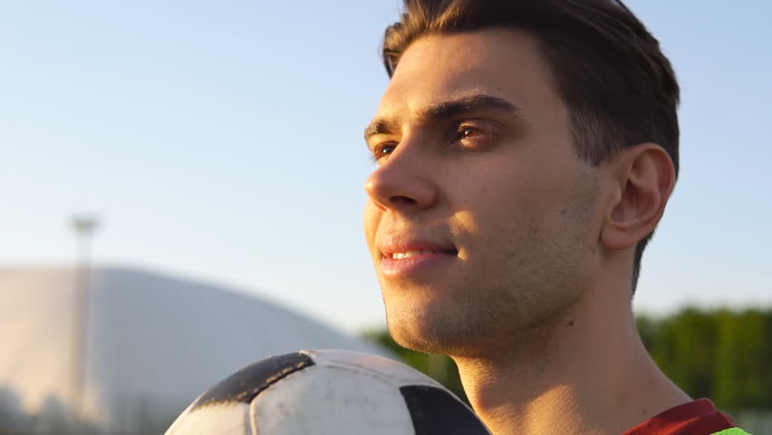 Close up portrait of a confident male football player with soccer ball standing at the stadium and looking away. Dreams come true.