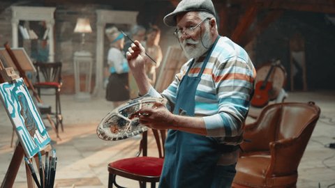 Slow motion footage of an old man dancing with a paint brush and palette in front of a canvas in an art studio Video de stock