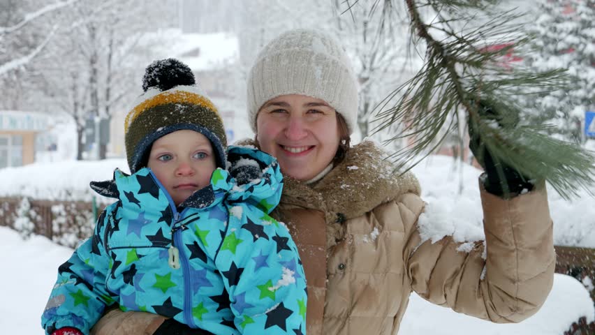 Portrait of happy smiling toddler boy with mother enjoying falling snow from tree branch at park. Fun and joy on winter holidays, kids playing outdoors, activity in snowfall