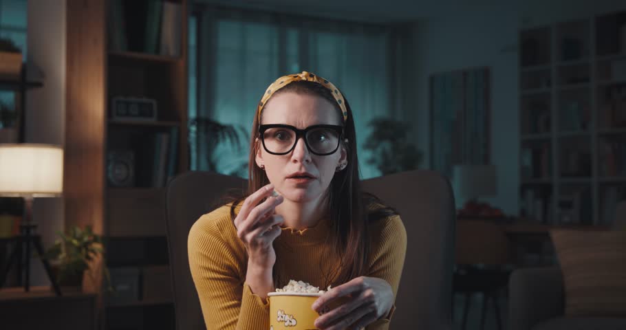 Shocked woman sitting on the armchair at home, she is watching action movies and eating popcorn Royalty-Free Stock Footage #1103810847