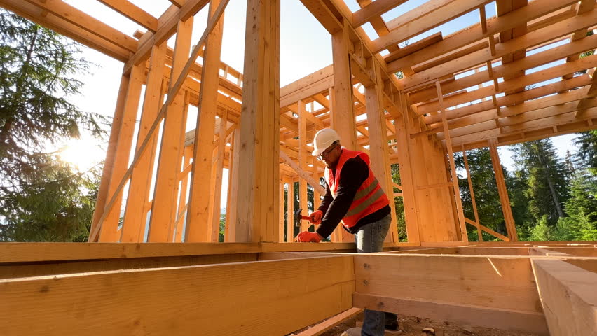 Carpenter constructing wooden frame house. Bearded man with glasses is hammering nails into the structure, wearing protective helmet and construction vest. Concept of modern ecological construction.