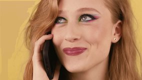 Smiling red-haired woman talking on the phone standing in front of the camera. A young lady standing on an orange background is having a pleasant conversation with her friend on the phone.