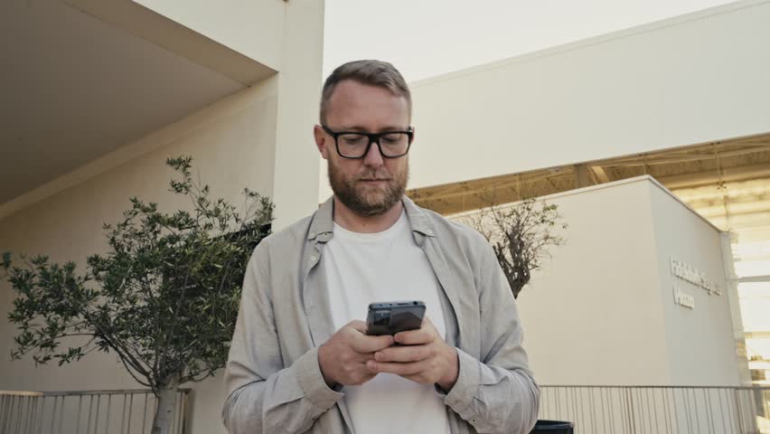 Attractive caucasian man in casual wear, stands outdoors, uses his smartphone, writes a message, texts with friends in social media, searches for information, typing email, looks phone screen, smiling