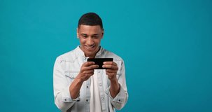 having fun with online gameplay - Young african american gamer playing mobile video games on smartphone standing over blue studio background