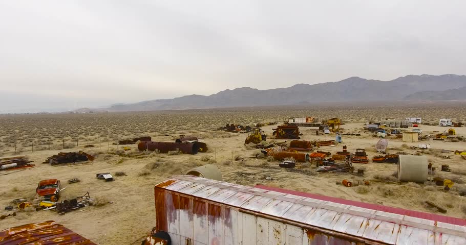 Aerial footage shot over a junk yard nestled in the Mojave desert. As the late morning sun struggles to break through the heavy overcast sky, the industrial area comes to life. 