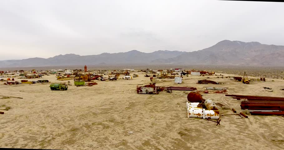 Aerial footage shot over a junk yard nestled in the Mojave desert. As the late morning sun struggles to break through the heavy overcast sky, the industrial area comes to life. 