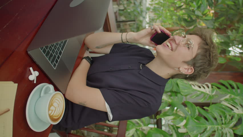 Vertical shot of young short-haired businesswoman sitting at table with cappuccino and laptop in outdoor cafe and speaking on cell phone