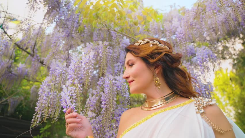 Fantasy woman greek goddess walks in spring garden hand touches purple wisteria flowers green tree summer nature day sun light Sexy girl queen old antique style white silk dress gold diadem jewelry 4k