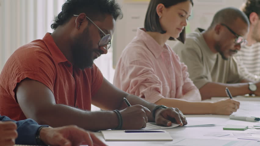 Medium portrait shot of multiracial man sitting at desk with diverse group mates and posing on camera while studying in school for immigrants