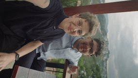 Vertical POV shot of young happy couple sitting in cafe with mountain view, waving and chatting on camera via online video call