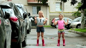 Two little girls in pink rubber boots are joyfully jumping in a rain puddle after a rainstorm in the courtyard of a high-rise building among cars. Slow motion. 4k video.