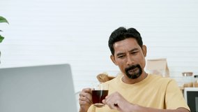 Close-up Asian man's eyes sitting on laptop computer, in living room at home, sit watching fun clips in online apps, on lazy days off from work, holding coffee mug in hand drink coffee.